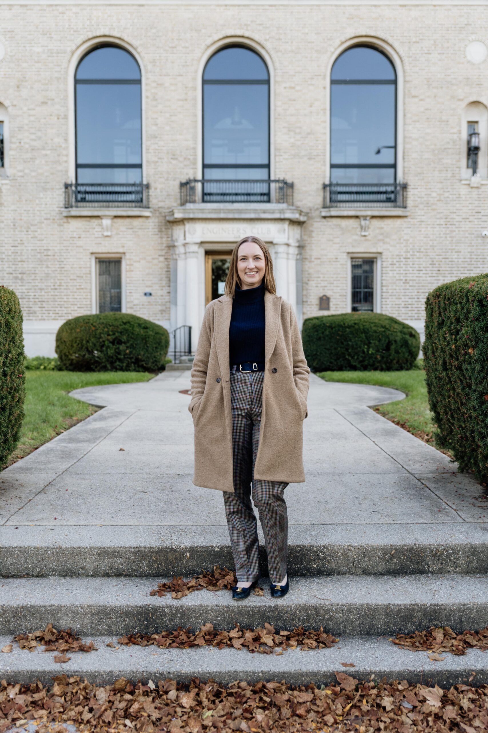 Author in beige coat and navy turtleneck standing in front of historic brick building.